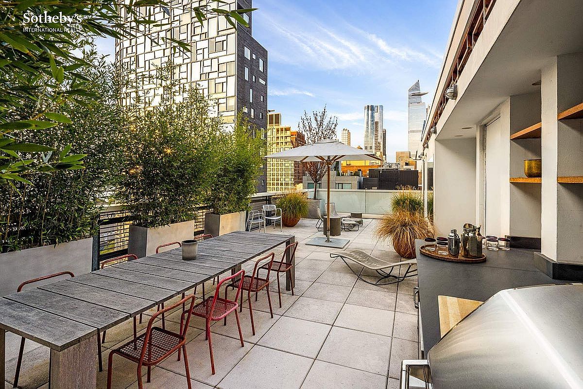 This expansive urban rooftop terrace offers a sophisticated outdoor living space with a long wooden dining table, modern red metal chairs, and a built-in outdoor kitchen area. The deck is framed by lush bamboo planters and features a lounge area with an umbrella, all set against a stunning backdrop of the city skyline. The perspective is a wide-angle shot from the terrace edge, emphasizing the seamless blend of luxury entertaining space and metropolitan views.