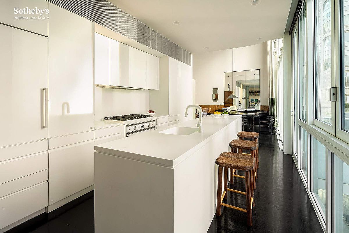 This sleek, minimalist kitchen features a long white island with a built-in sink and three woven bar stools, set against a backdrop of high-gloss white cabinetry and dark hardwood flooring. A wall of floor-to-ceiling windows floods the space with natural light, while a large mirror at the far end enhances the sense of depth and openness. The design is ultra-modern and sophisticated, emphasizing clean lines and a bright, airy atmosphere.