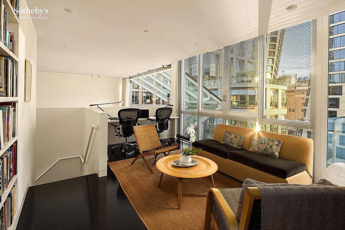 This bright, modern home office and lounge area features a sleek desk setup with dual ergonomic chairs, a comfortable sofa, and a woven accent chair arranged around a circular coffee table. The space is defined by floor-to-ceiling windows that flood the room with natural light, complemented by dark hardwood floors and a built-in bookshelf. The perspective is a wide-angle shot from the corner of the room, capturing the seamless blend of a productive workspace and a relaxing living area.