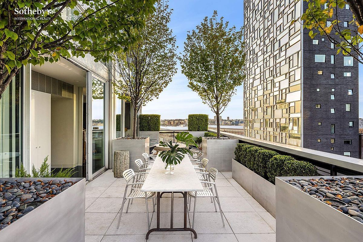 This sophisticated urban terrace features a long, elegant dining table set for outdoor entertaining, surrounded by manicured boxwood hedges and sculptural trees in modern planters. The space offers a stunning, elevated view of the river and city skyline, framed by contemporary architecture and large glass sliding doors. The overall impression is one of luxury, tranquility, and high-end metropolitan living.