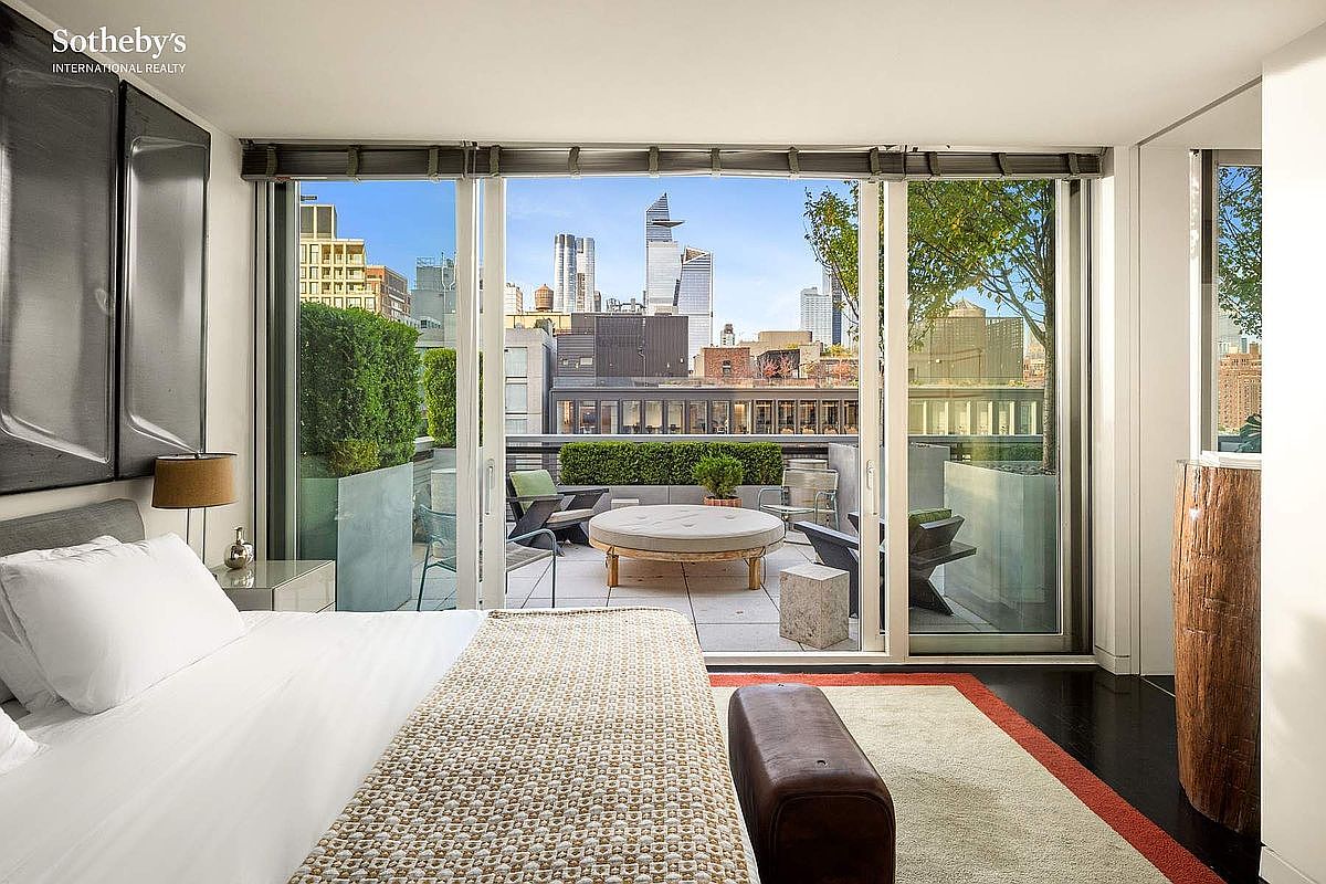 This sophisticated primary bedroom features a plush bed with crisp white linens and a textured throw, leading directly to a private terrace through expansive floor-to-ceiling sliding glass doors. The room is anchored by a dark wood floor and a neutral area rug, while the outdoor space offers a serene urban oasis with a circular lounge chair and lush greenery against a stunning city skyline backdrop. The overall aesthetic is one of modern luxury, seamlessly blending indoor comfort with high-end outdoor living.