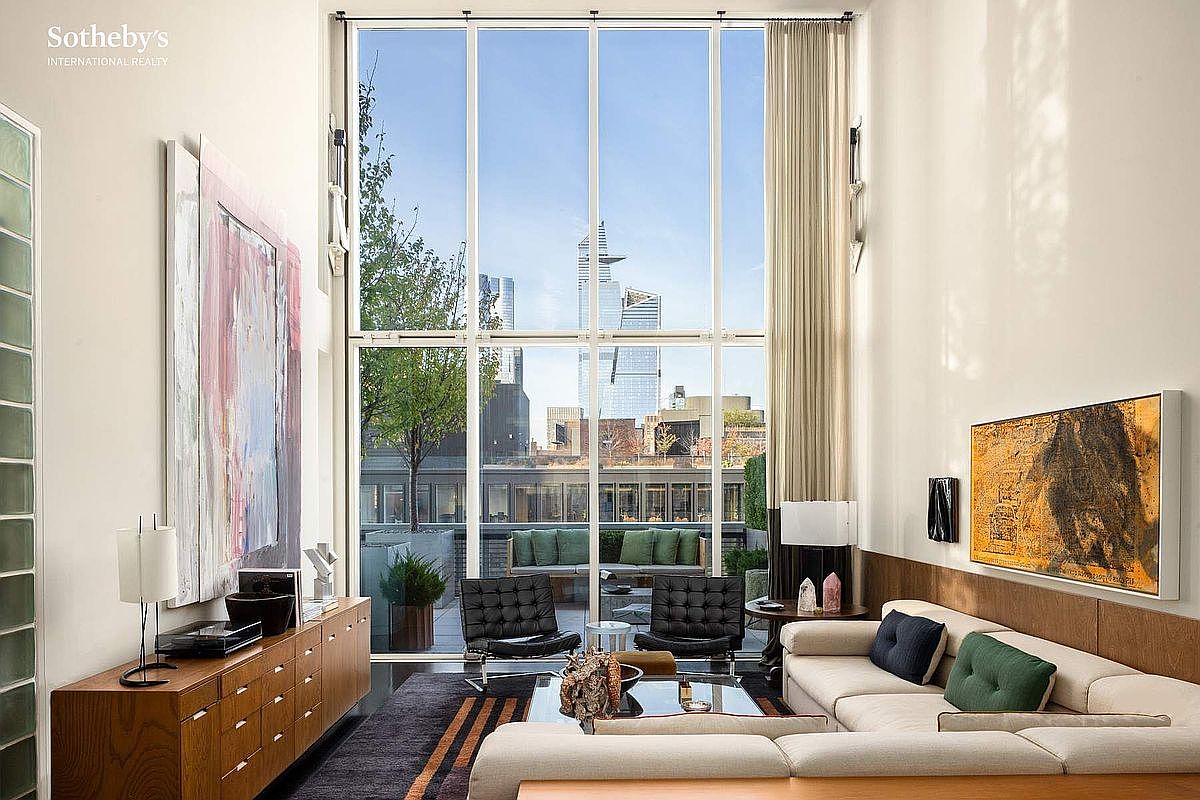 This sophisticated, double-height living room features floor-to-ceiling windows that frame a stunning urban skyline view and open onto a private terrace. The space is elegantly furnished with a plush sectional sofa, iconic mid-century modern lounge chairs, and a sleek wooden credenza, all complemented by large-scale contemporary artwork. The high ceilings and abundant natural light create an airy, luxurious atmosphere perfect for high-end city living.