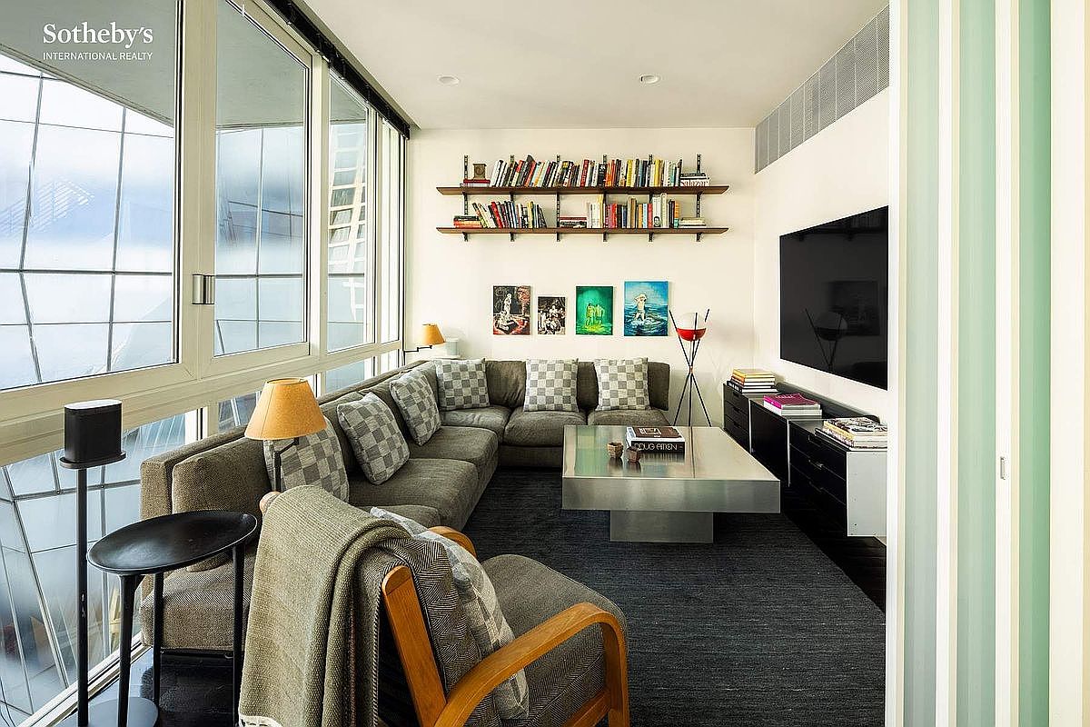 This modern, sophisticated living room features a large, plush sectional sofa arranged in a U-shape, centered around a sleek, metallic coffee table. The space is accented by a wall of floating bookshelves, a large wall-mounted television, and a series of colorful art pieces, all illuminated by natural light streaming through expansive floor-to-ceiling windows. The overall aesthetic is contemporary and cozy, with a neutral color palette and a comfortable, inviting atmosphere.