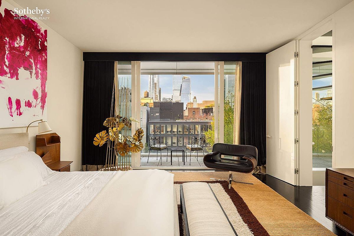 This sophisticated primary bedroom features a minimalist aesthetic with a large, plush bed and a striking abstract art piece on the wall. A floor-to-ceiling glass door opens onto a private balcony, offering a stunning urban view of the city skyline. The room is accented by a sleek black lounge chair, a textured area rug, and warm, ambient lighting, creating a luxurious and serene atmosphere.