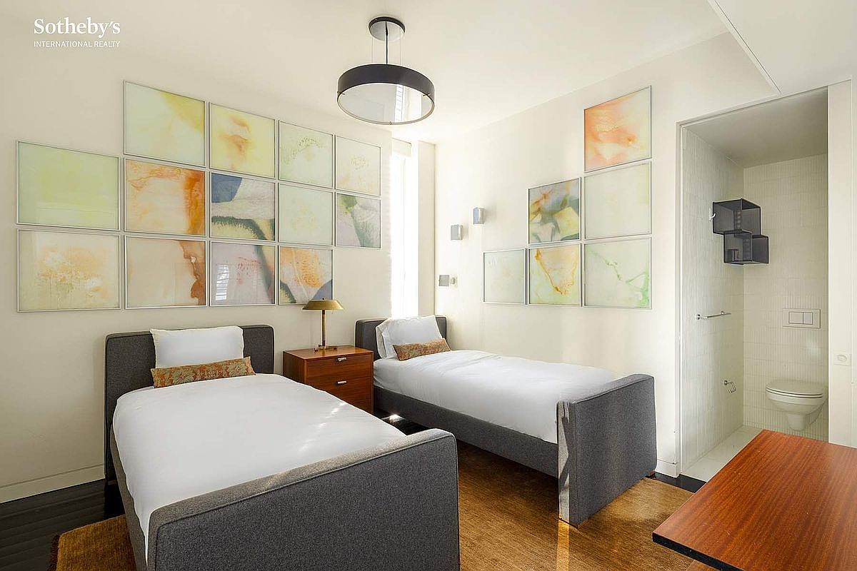 This bright and modern guest bedroom features two twin beds with grey upholstered frames, separated by a warm wood nightstand. The walls are adorned with a grid of abstract, soft-toned artwork, and the room includes an en-suite bathroom visible through an open doorway. The space is illuminated by a contemporary pendant light and natural light, creating a clean, inviting, and well-organized atmosphere.