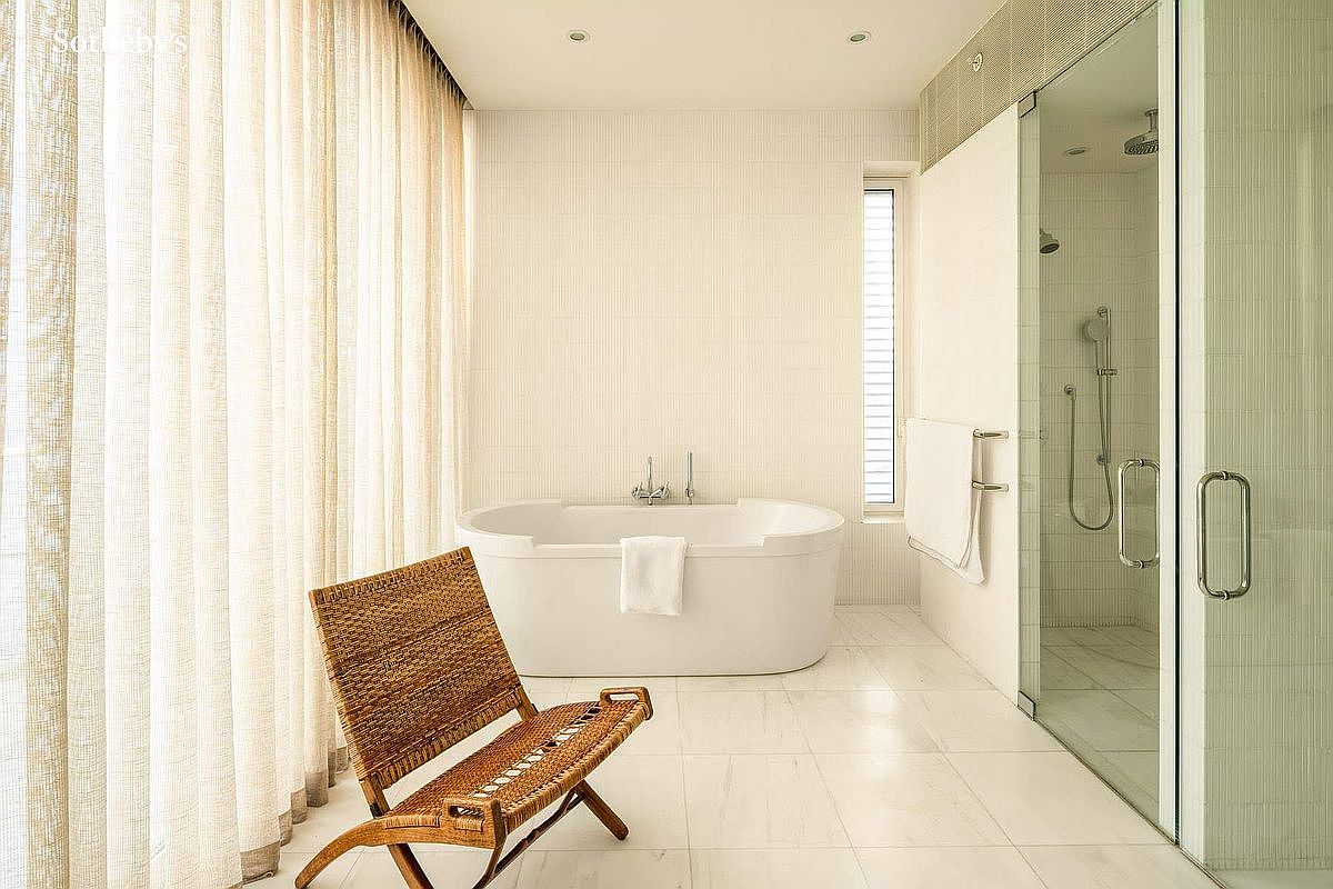This bright and airy primary bathroom features a sleek, freestanding soaking tub as its centerpiece, complemented by a large glass-enclosed walk-in shower. A woven wooden chair sits in the foreground, adding a touch of organic texture to the minimalist, light-toned space. The room is bathed in soft, natural light filtered through floor-to-ceiling sheer curtains, creating a serene and spa-like atmosphere.