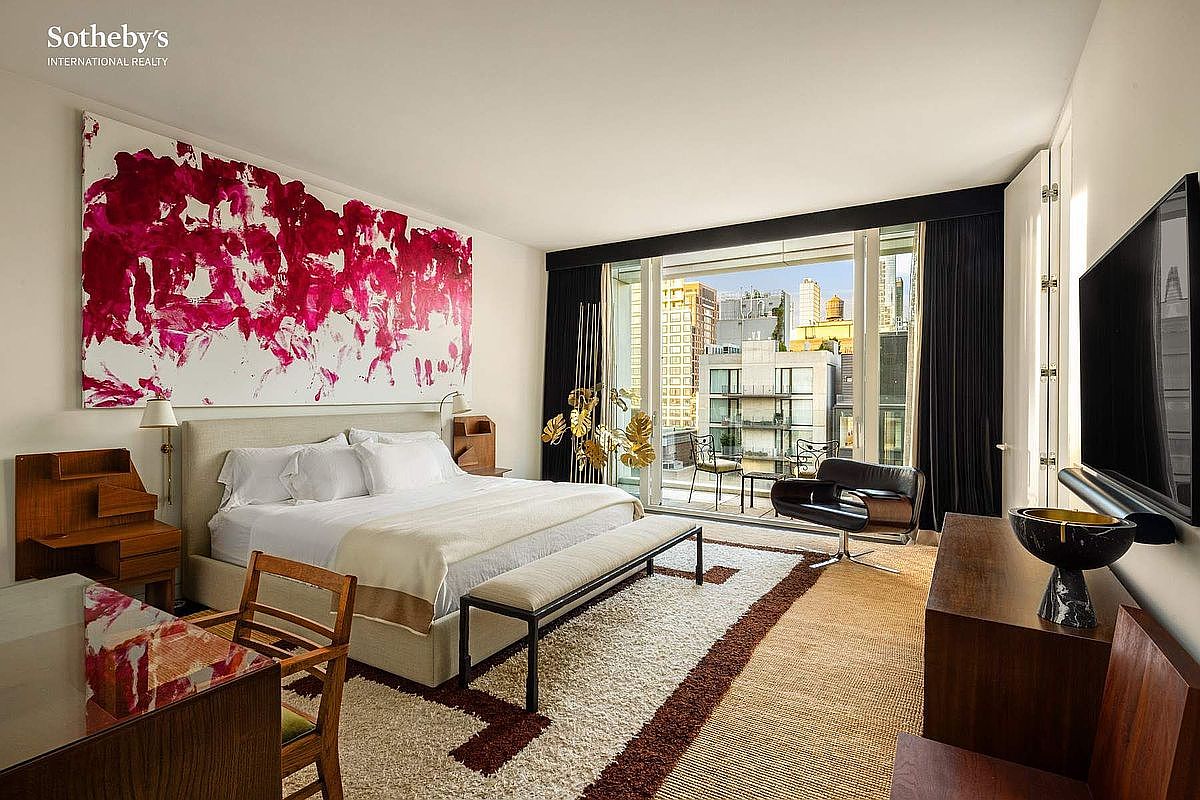 This sophisticated primary bedroom features a large, plush bed centered beneath a striking, oversized abstract painting with vibrant magenta accents. The room is bathed in natural light from floor-to-ceiling windows that open onto a private balcony, offering an urban view. A textured area rug, a sleek modern armchair, and warm wood furnishings create a luxurious and contemporary atmosphere.
