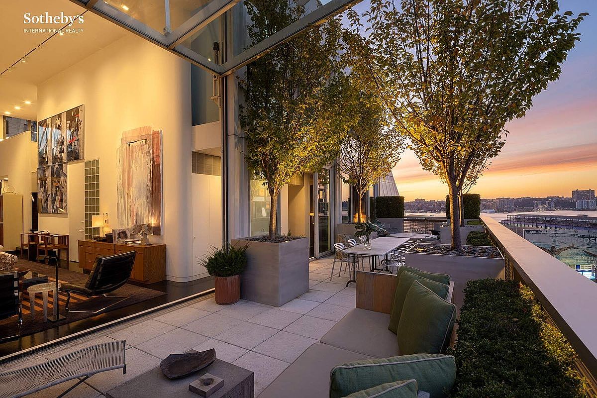 This luxurious high-rise terrace offers a seamless indoor-outdoor living experience, featuring expansive glass walls that open onto a spacious, paved balcony. The space is elegantly furnished with modern outdoor seating and dining areas, complemented by mature trees in sleek planters and a stunning sunset view over the city skyline and waterfront. The overall atmosphere is sophisticated and serene, highlighting a premium urban lifestyle.