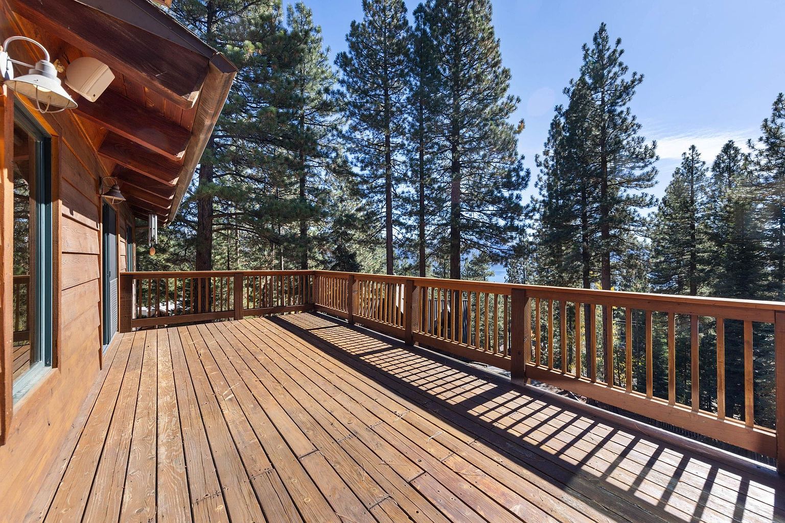 This image showcases a spacious wooden deck attached to a house, surrounded by tall pine trees. The deck features a wooden railing and ample space for outdoor furniture. The warm tones of the wood contrast with the green of the trees, creating a serene and inviting outdoor living space.