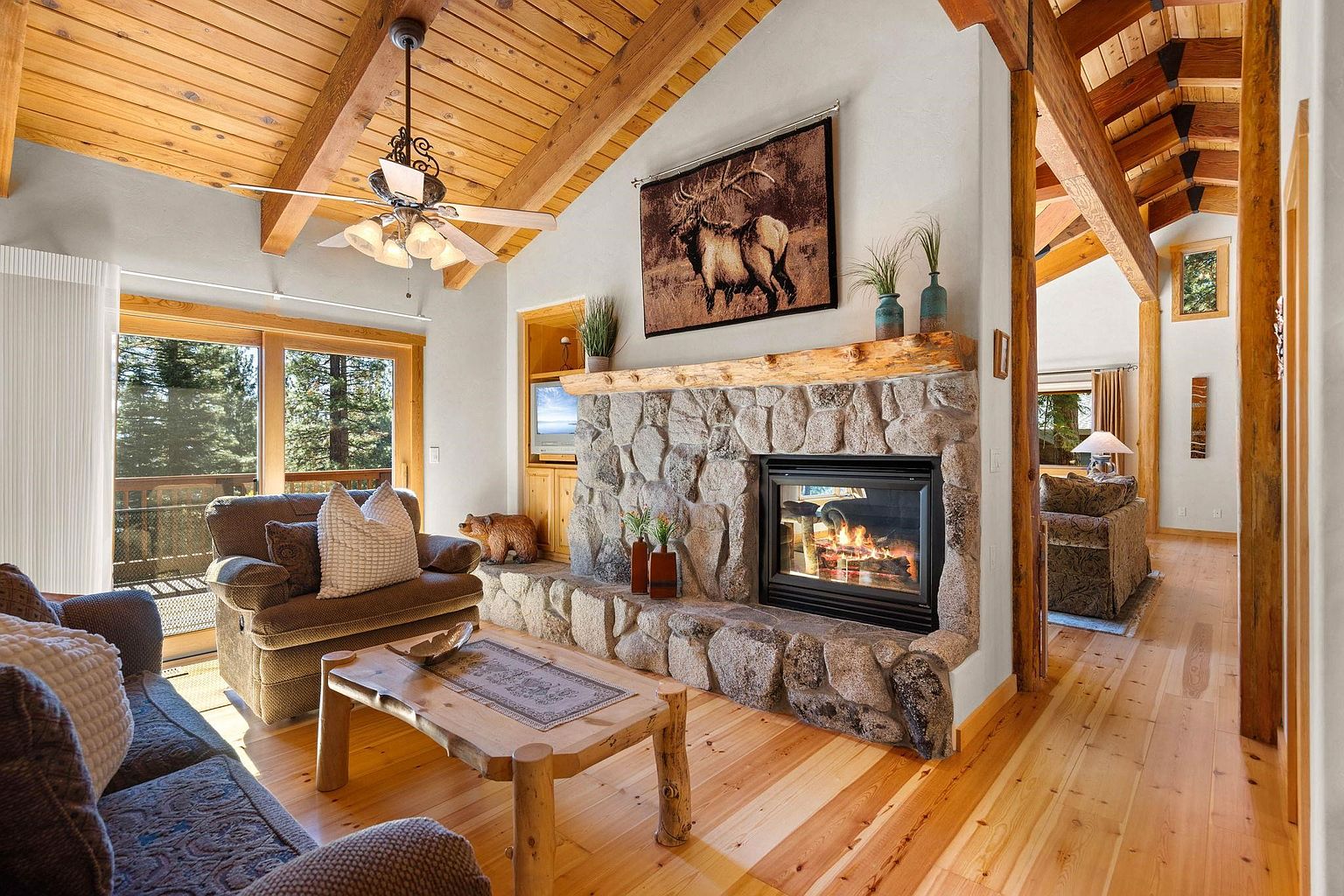 This inviting living room features a stone fireplace with a wooden mantel, complemented by a tapestry of elk above. The room is furnished with comfortable sofas and a rustic wooden coffee table, set against a backdrop of wood-paneled ceilings and hardwood floors. A sliding glass door leads to an outdoor balcony, offering natural light and views of the surrounding trees.