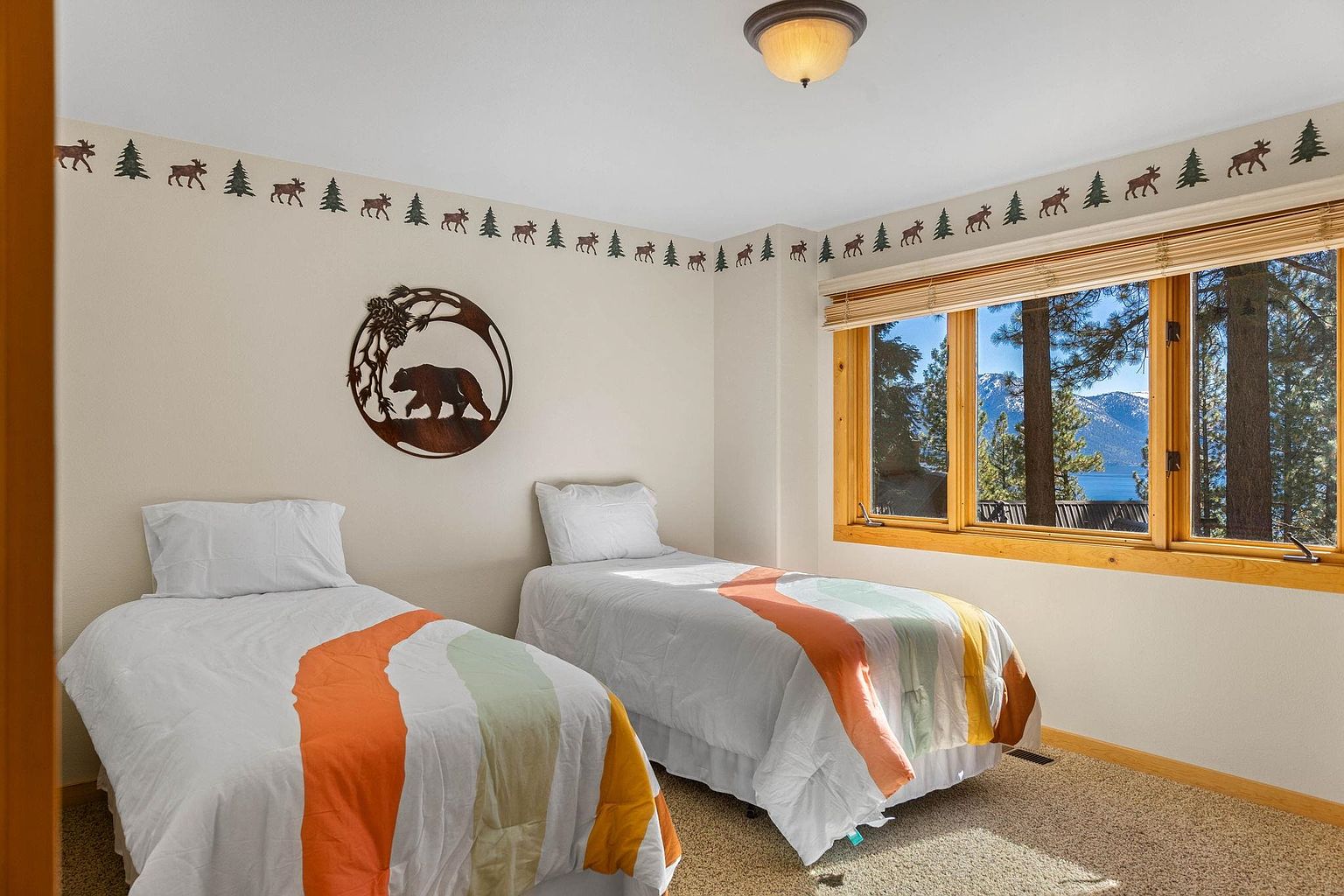 This is a cozy guest bedroom featuring two twin beds with white linens and colorful striped throws. A wooden-framed window offers a view of the surrounding trees and a glimpse of a lake. The room is decorated with a nature-themed border and a metal bear wall art, creating a warm and inviting atmosphere.