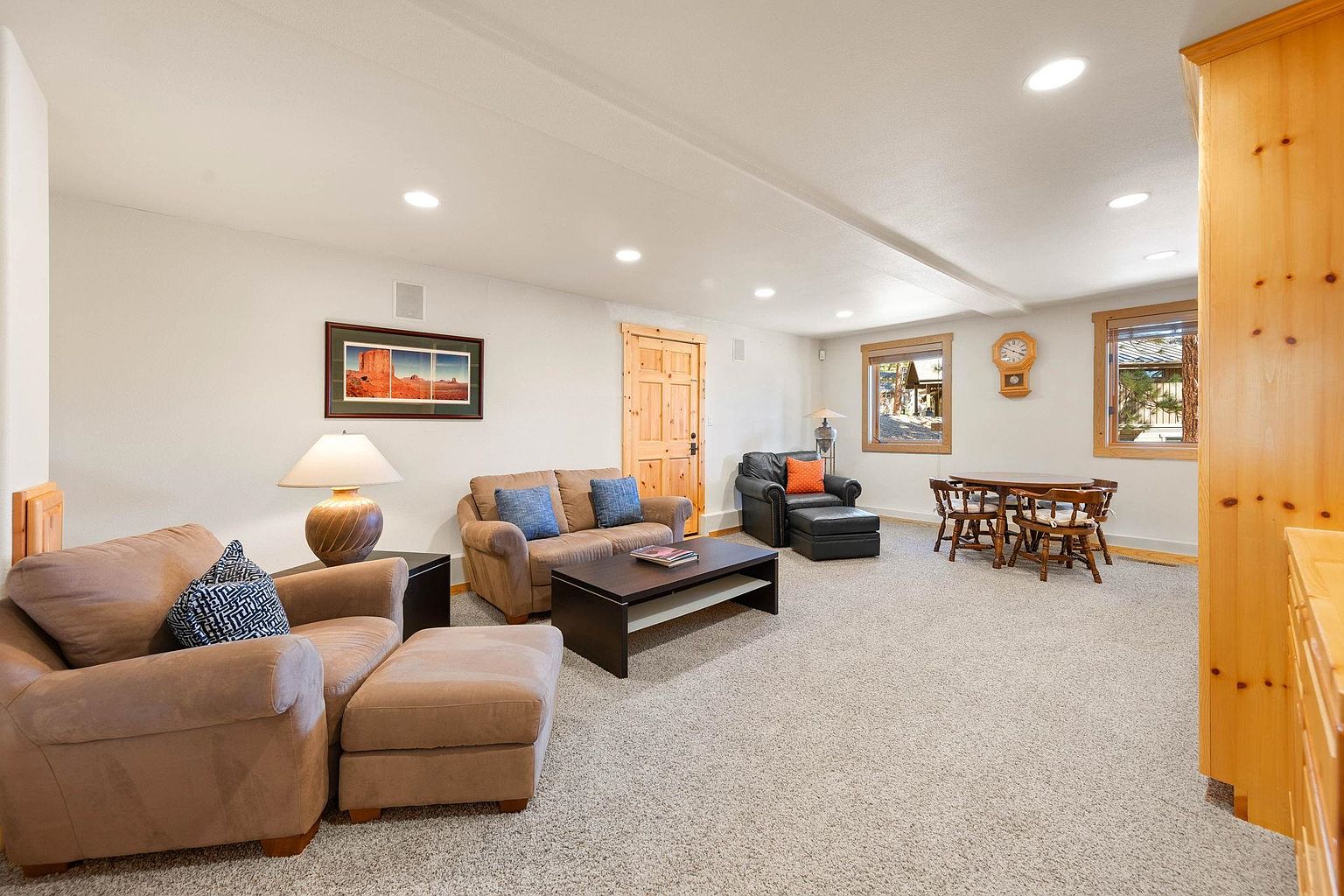 This is an interior shot of a cozy living room featuring a brown sofa and armchair with decorative pillows, a dark wood coffee table, and a black leather chair with ottoman. The room is well-lit with recessed lighting and natural light from the windows, which are complemented by a wooden dining table set. The overall impression is warm and inviting, with a neutral color palette and comfortable furnishings.
