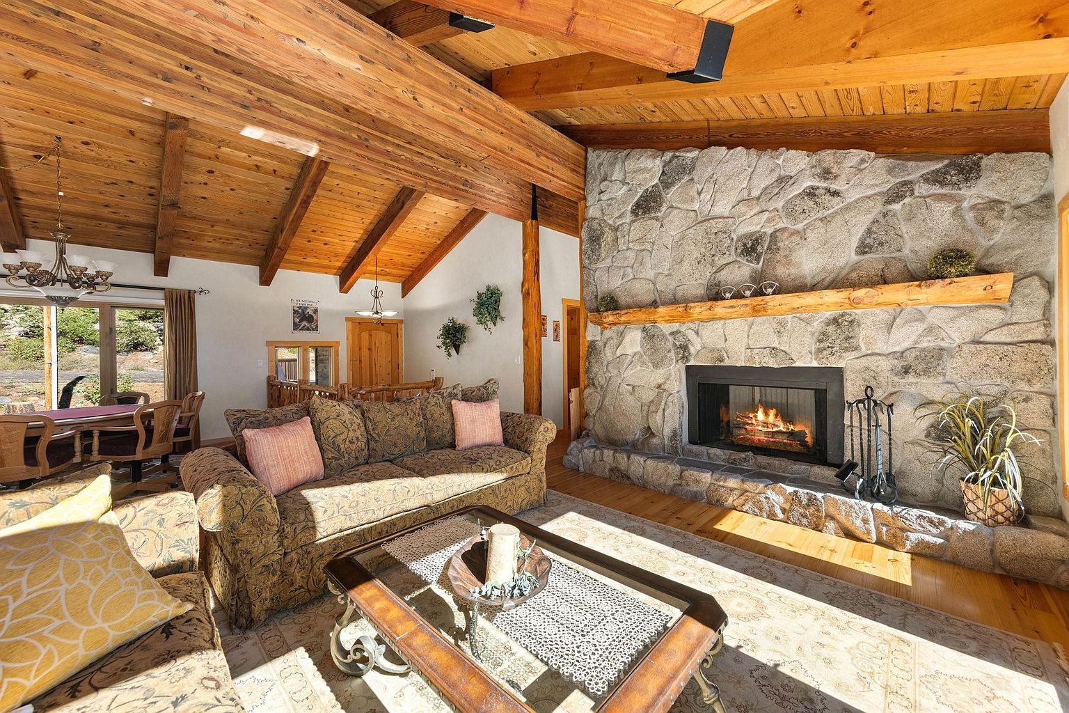 This inviting living room features a stone fireplace as a focal point, complemented by warm wood ceilings and beams. A comfortable patterned sofa with decorative pillows sits atop a patterned rug, creating a cozy and stylish atmosphere. The room is well-lit, showcasing the textures and colors of the furnishings and architectural details.