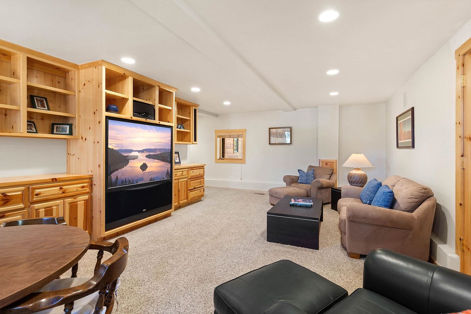 This is an interior shot of a cozy living room featuring built-in wooden shelving and cabinetry that houses a large television. The room is furnished with comfortable brown sofas, a black coffee table, and a black leather ottoman, all set on a neutral-toned carpet. The lighting is provided by recessed ceiling lights and a table lamp, creating a warm and inviting atmosphere.