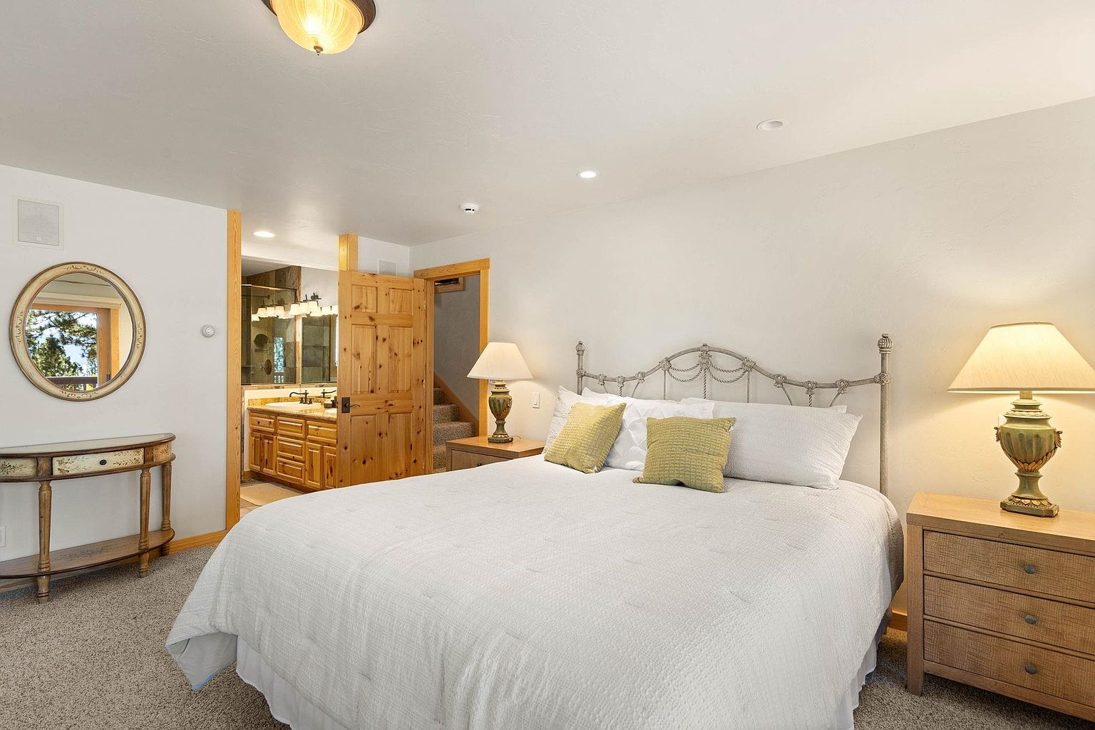This is a well-lit primary bedroom featuring a wrought iron bed frame with white bedding and decorative pillows. A wooden nightstand with a lamp sits beside the bed, and a round mirror hangs above a decorative table on the opposite wall. The room has a cozy and inviting atmosphere, with a glimpse into an adjacent bathroom.
