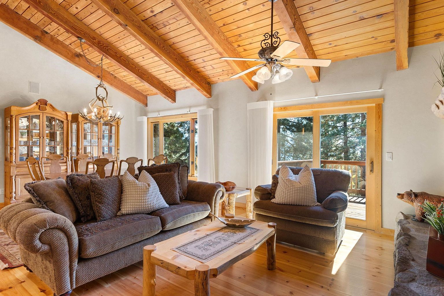 This is an interior shot of a cozy living room featuring a brown sofa and armchair, a wooden coffee table, and a large wooden cabinet in the background. The room has a high, wood-paneled ceiling with exposed beams and a ceiling fan. Large windows provide natural light and views of the outdoors, creating a warm and inviting atmosphere.
