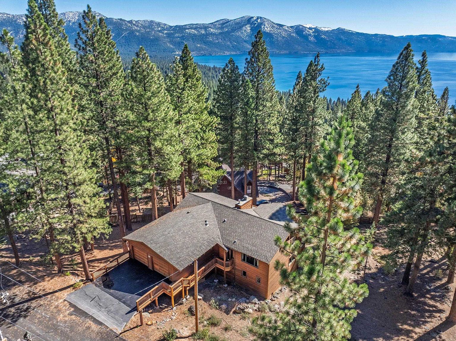 This aerial shot showcases a beautiful wooden house nestled among tall pine trees, with a stunning view of a lake and snow-capped mountains in the background. The house features a gray shingle roof, wooden decks, and a paved driveway, blending harmoniously with the natural surroundings. The overall impression is one of serene luxury and privacy.