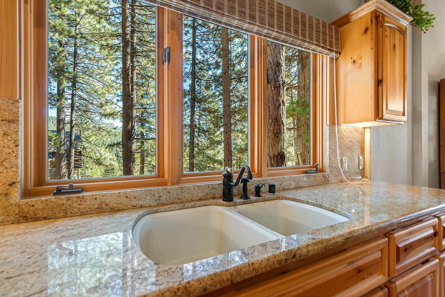This image showcases a kitchen featuring a double basin sink with a dark faucet set against a backdrop of a large window offering a view of a dense forest. The countertops are a speckled granite, and the cabinetry is a warm wood tone, creating a cozy and natural atmosphere. A wooden cabinet is visible on the right.