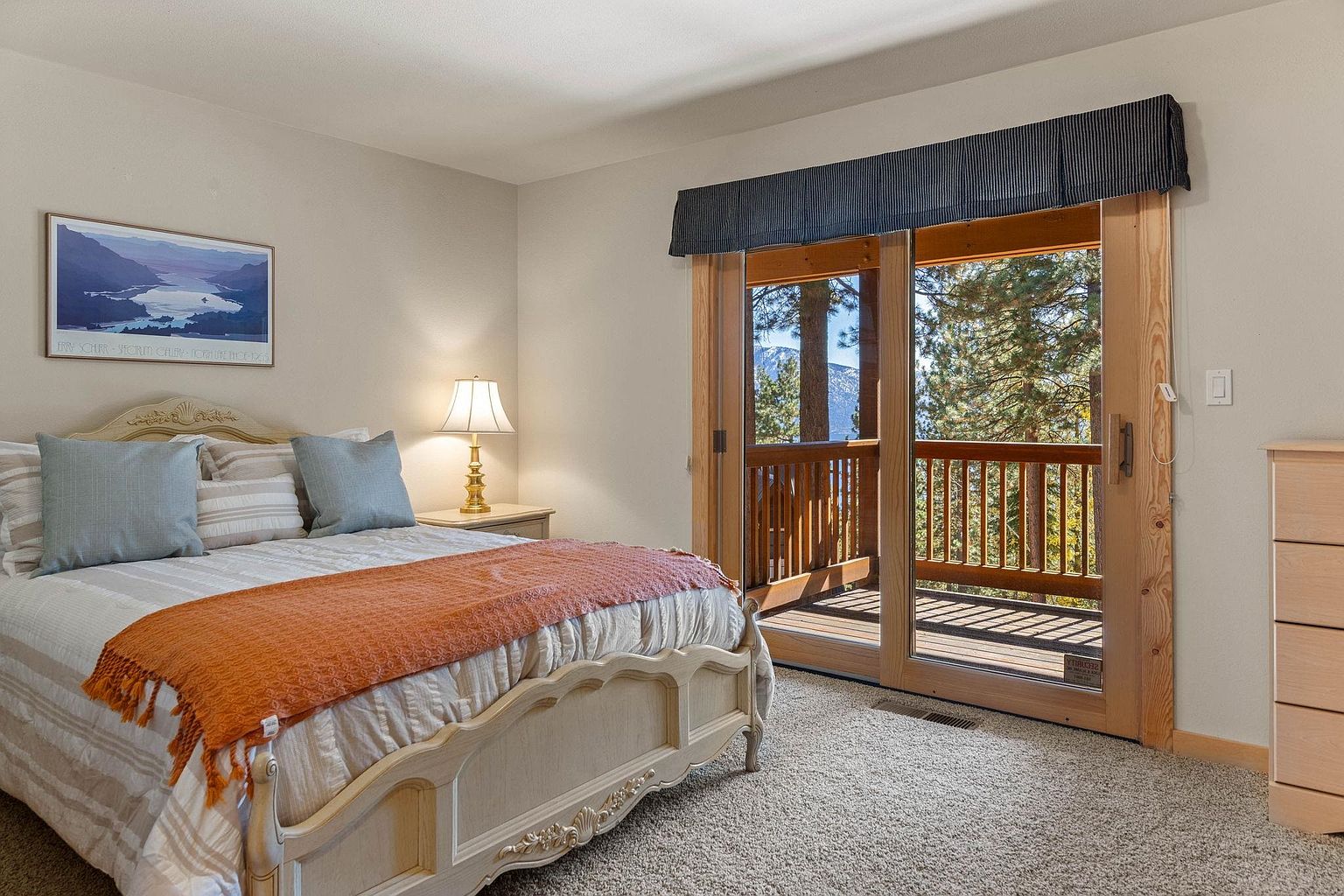 This is a cozy primary bedroom featuring a decorative bed frame with an orange throw blanket, two blue pillows, and striped bedding. A bedside table with a lamp sits next to the bed, and sliding glass doors lead to a balcony with a view of trees and mountains. The room has neutral-toned walls and carpet, creating a warm and inviting atmosphere.