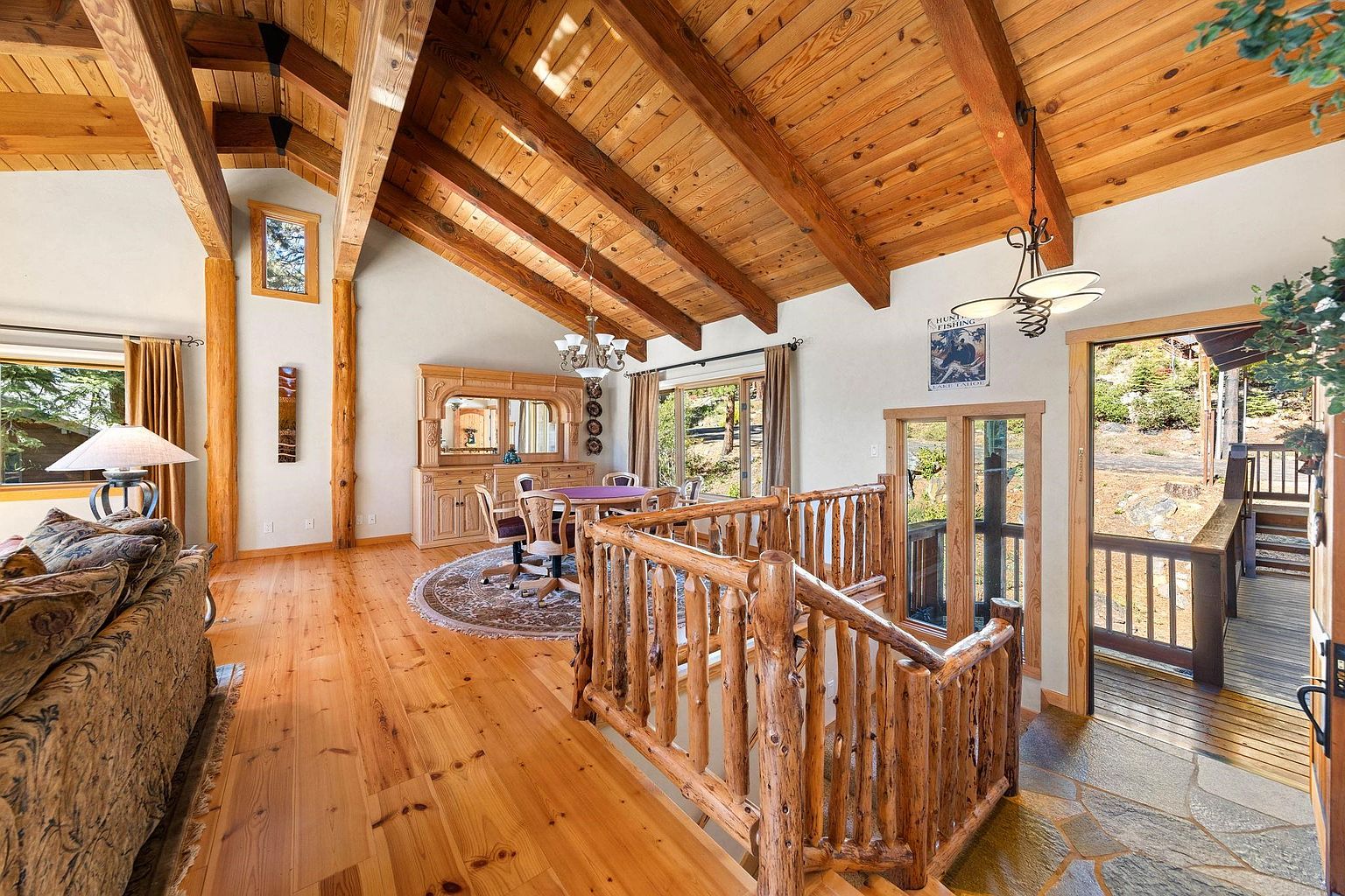 This interior shot showcases a spacious living area with a rustic, lodge-like aesthetic. The room features exposed wooden beams, hardwood floors, and a unique log railing leading to another level. A dining area with a wooden table and chairs is visible in the background, along with a large wooden cabinet and a view to the outside through a set of doors.