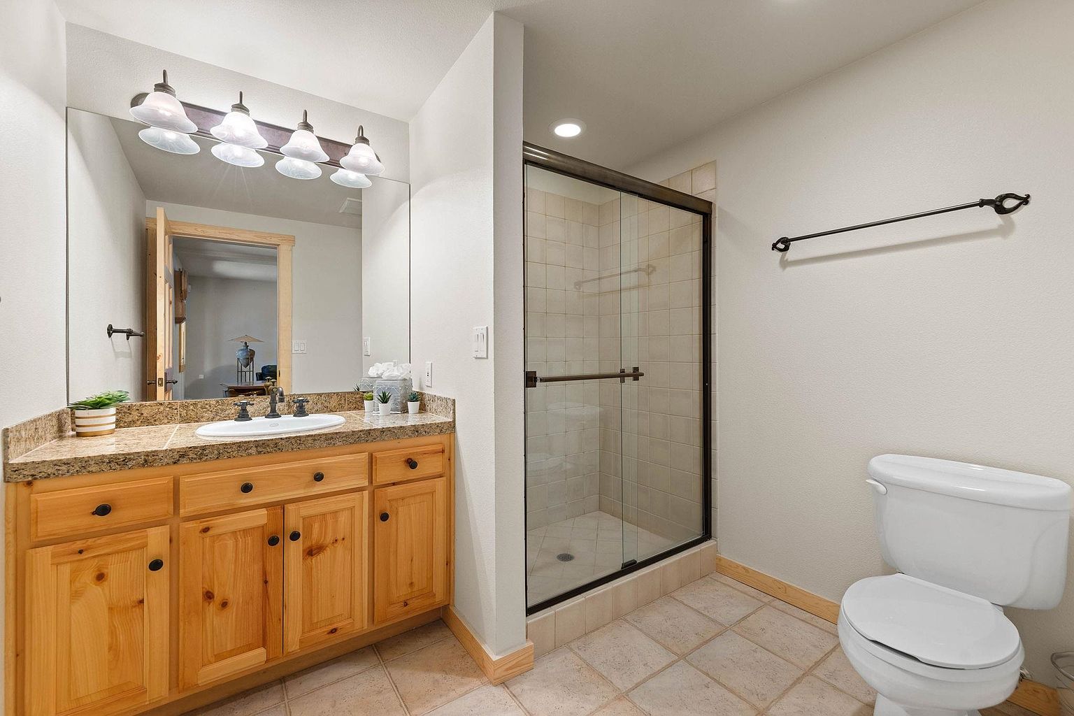 This is a well-lit bathroom featuring a wooden vanity with a granite countertop, a white sink, and a large mirror with decorative lighting. A glass-enclosed shower is adjacent to the vanity, and a white toilet is positioned on the right side of the frame. The floor is tiled, and the overall impression is clean and functional.