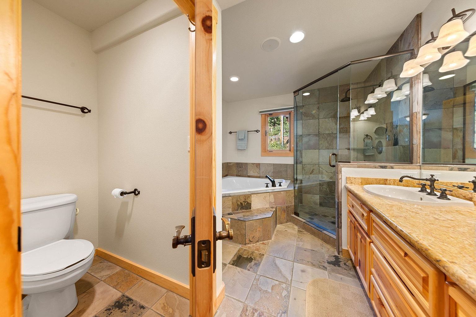 This is a primary bathroom featuring a toilet, a soaking tub, a glass-enclosed shower, and a vanity with a granite countertop. The bathroom has a rustic style with stone tile flooring and wood trim. The overall impression is luxurious and well-appointed.