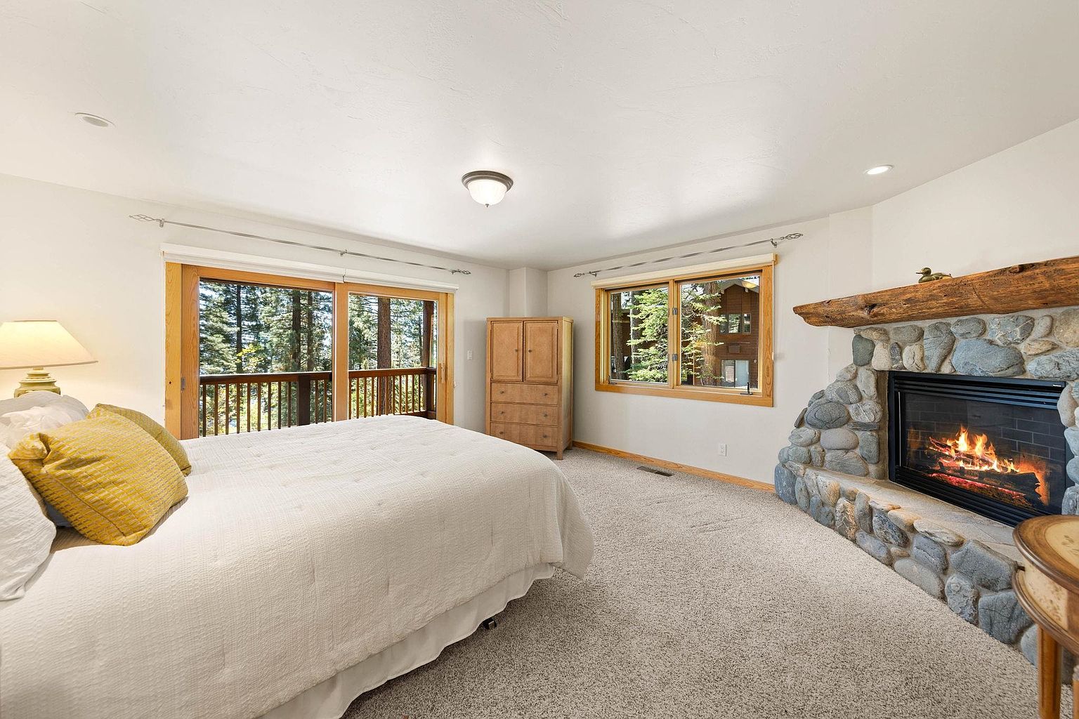 This is a primary bedroom featuring a large bed with white linens and yellow pillows, a stone fireplace with a wooden mantel, and large windows offering views of the surrounding trees. A wooden armoire provides storage, and the room is carpeted in a neutral tone. The overall impression is cozy and inviting, with a focus on natural light and rustic elements.