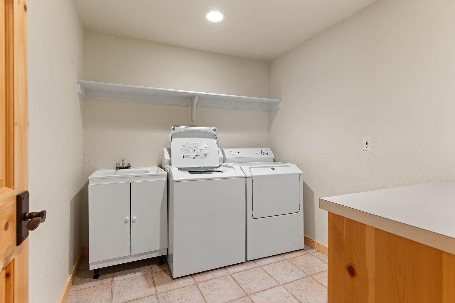 This is a well-lit laundry room featuring a white washer and dryer set, a utility sink with a white cabinet, and a shelf above. The room has neutral-colored walls and tiled flooring. A wooden cabinet is partially visible on the right side of the frame, and a wooden door is on the left, adding a touch of warmth to the space.