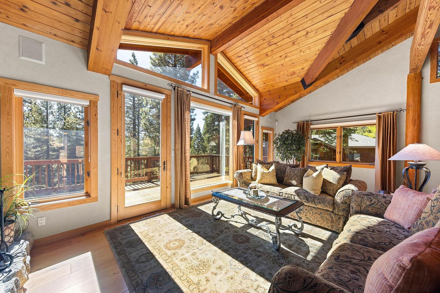 This inviting living room features a high, wood-paneled ceiling with exposed beams, creating a warm and rustic atmosphere. Large windows and glass doors offer abundant natural light and views of the surrounding trees and deck. The room is furnished with a comfortable, patterned sofa and a decorative rug, enhancing the cozy and welcoming feel of the space.