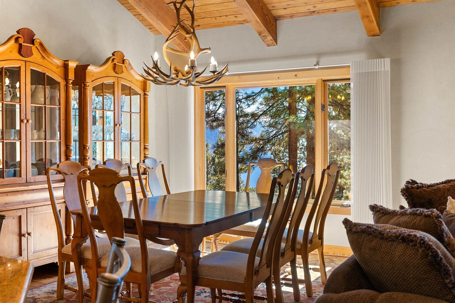 This is an interior shot of a dining room featuring a wooden dining table surrounded by wooden chairs with upholstered seats. A large wooden hutch stands against the wall, and a unique antler chandelier hangs above the table. A large window offers a view of the outdoors, bringing natural light into the space.