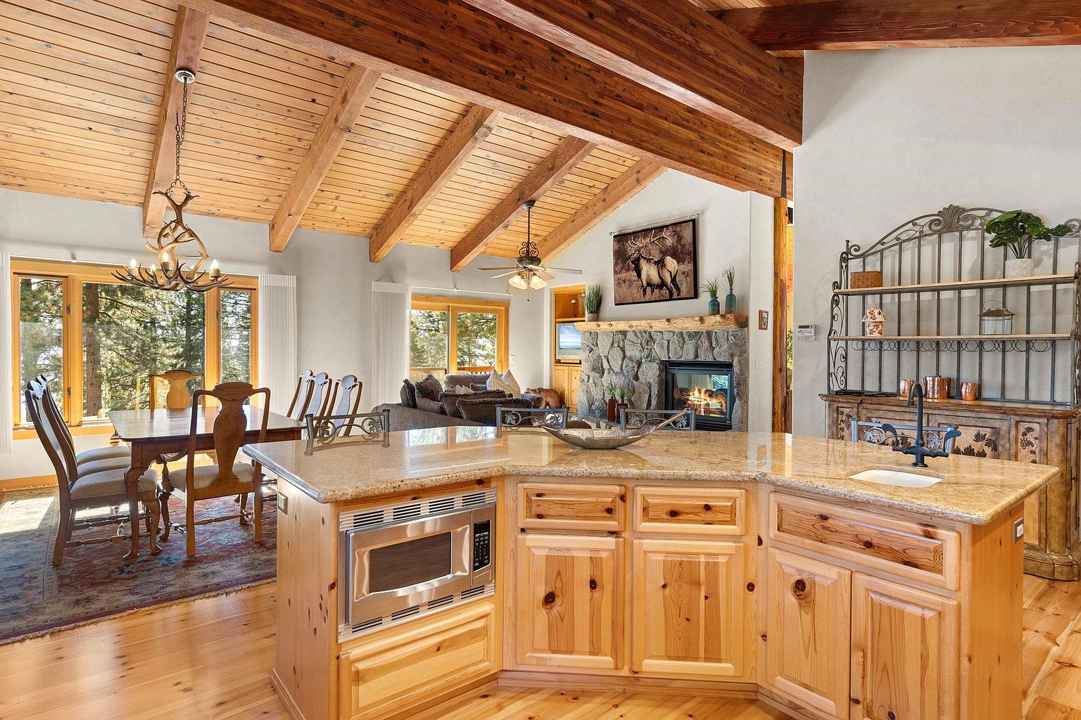 This interior shot showcases a warm and inviting kitchen that seamlessly blends into the dining and living areas. The kitchen features light wood cabinetry, granite countertops, and stainless steel appliances, while the dining area boasts a wooden table and chairs under a unique antler chandelier. The living area is visible in the background, complete with a stone fireplace and comfortable seating, all under a striking wooden beamed ceiling.