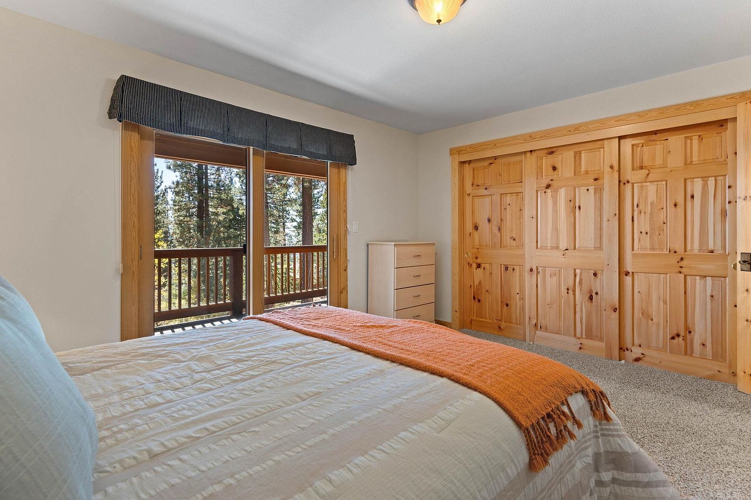 This is a cozy bedroom featuring a bed with a white comforter and an orange throw blanket. The room has a sliding door closet with wooden doors and a small dresser. A window with a balcony offers a view of the surrounding trees, creating a serene and inviting atmosphere.