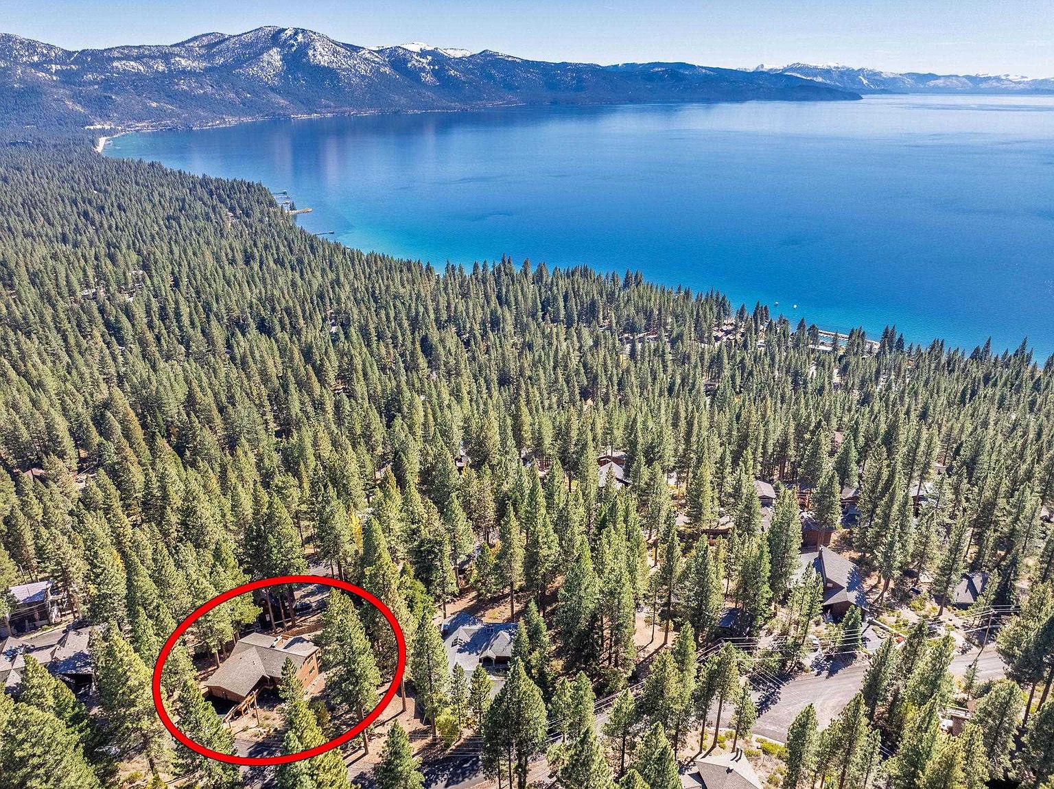 This aerial shot showcases a charming home nestled among towering evergreen trees, with a stunning view of a large, serene lake and snow-capped mountains in the background. The house, circled in red, features a brown roof and blends harmoniously with its natural surroundings, offering a sense of privacy and tranquility. The landscape emphasizes the property's secluded location and access to natural beauty.