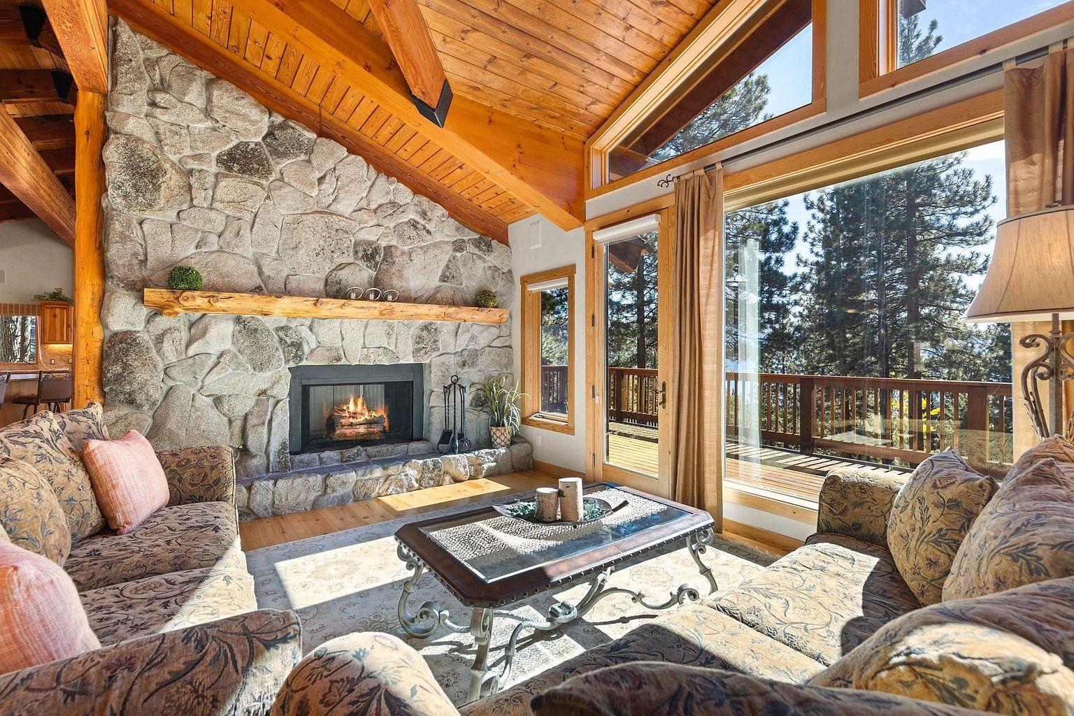 This is an inviting living room featuring a stone fireplace with a wooden mantel, creating a cozy focal point. The room is furnished with comfortable sofas and a decorative coffee table, complemented by natural light streaming through large windows that offer a view of the outdoors. The wooden ceiling adds warmth and character to the space, enhancing the overall rustic yet elegant ambiance.