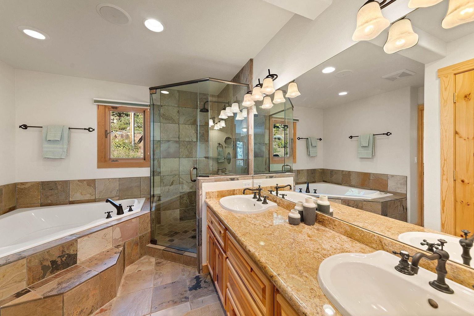This is a primary bathroom featuring a double vanity with granite countertops and wooden cabinets. A glass-enclosed shower and a separate bathtub with stone tile surround add to the luxurious feel. The room is well-lit with recessed lighting and decorative sconces, creating a warm and inviting atmosphere.