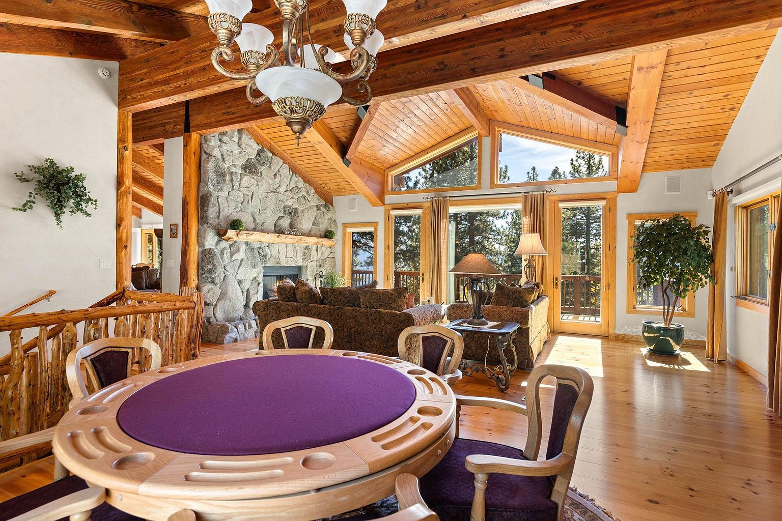 This is an interior shot of a spacious living room featuring a stone fireplace, wood-beamed ceilings, and large windows offering natural light. The room includes comfortable seating, a game table with chairs, and hardwood floors, creating a warm and inviting atmosphere. The overall style is rustic and luxurious, perfect for a mountain retreat.