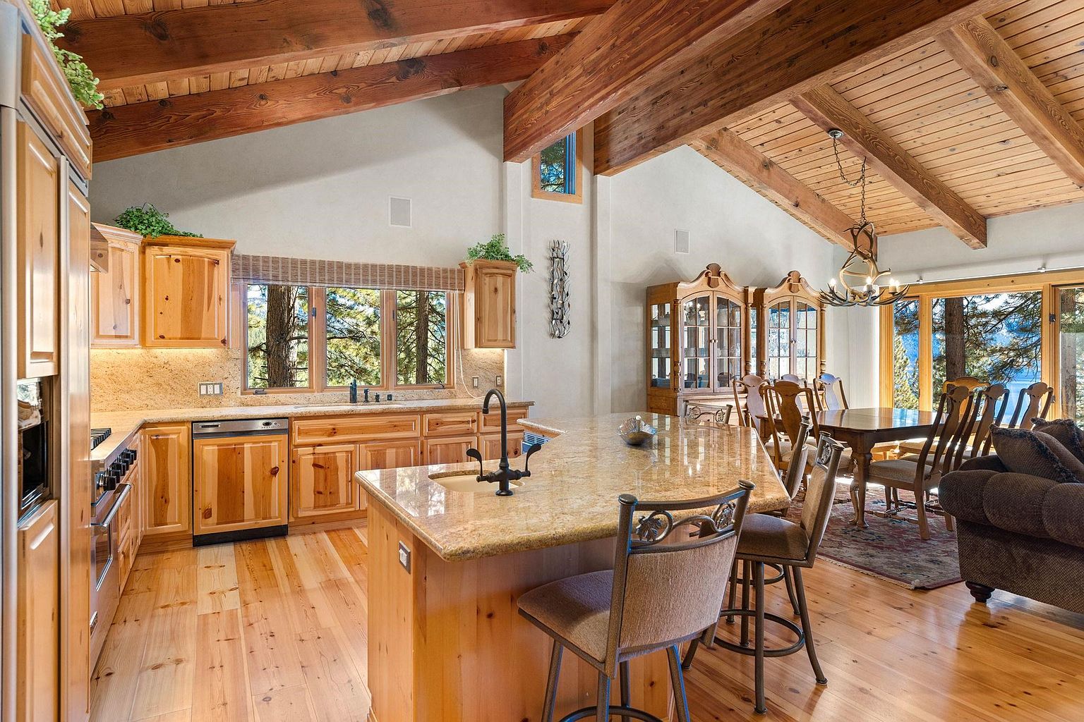 This is a warm and inviting kitchen and dining area featuring natural wood cabinetry, granite countertops, and hardwood floors. The kitchen island provides seating, and the dining area includes a wooden table and chairs. The space is well-lit with natural light from the windows and a decorative chandelier, creating a cozy and elegant atmosphere.