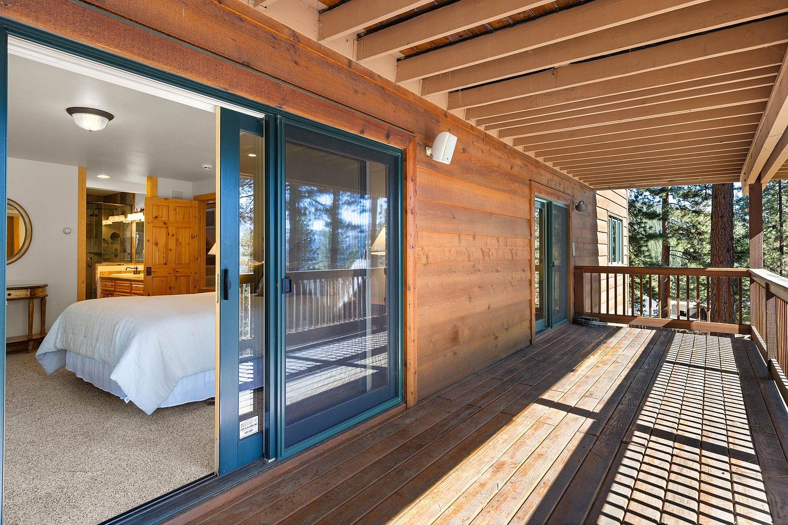 The image showcases a spacious wooden deck extending from a home, offering an inviting outdoor living space. A sliding glass door provides direct access to a bedroom, while the deck itself features wooden railings and ample sunlight. The view beyond the deck includes trees, suggesting a serene and private setting.