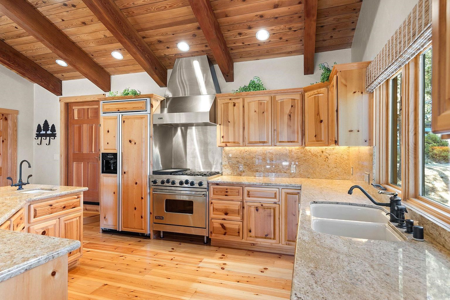 This is a well-lit kitchen featuring light wood cabinetry, granite countertops, and stainless steel appliances, including a Viking range and hood. The ceiling is wood-paneled with exposed beams, adding a rustic touch. A large window provides natural light and a view to the outside.