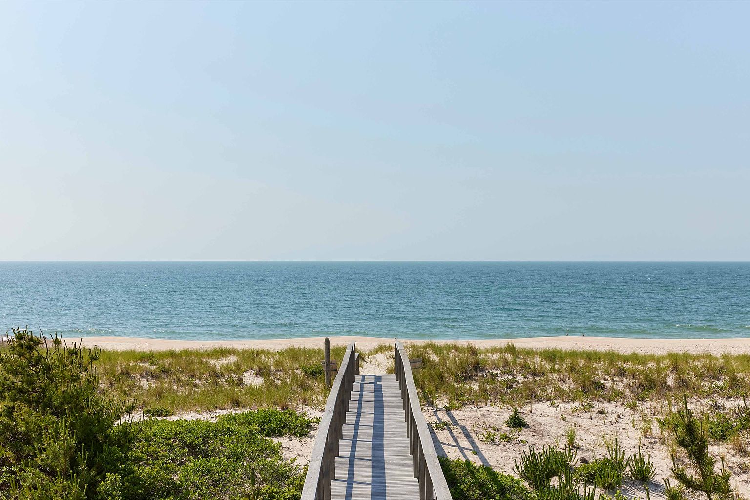 This image showcases a wooden boardwalk leading from a lush, green yard to a serene beach and ocean. The yard features a mix of low-lying shrubs and taller grasses, creating a natural and inviting landscape. The boardwalk provides direct access to the sandy beach and the calm, blue ocean, suggesting a private and tranquil setting.