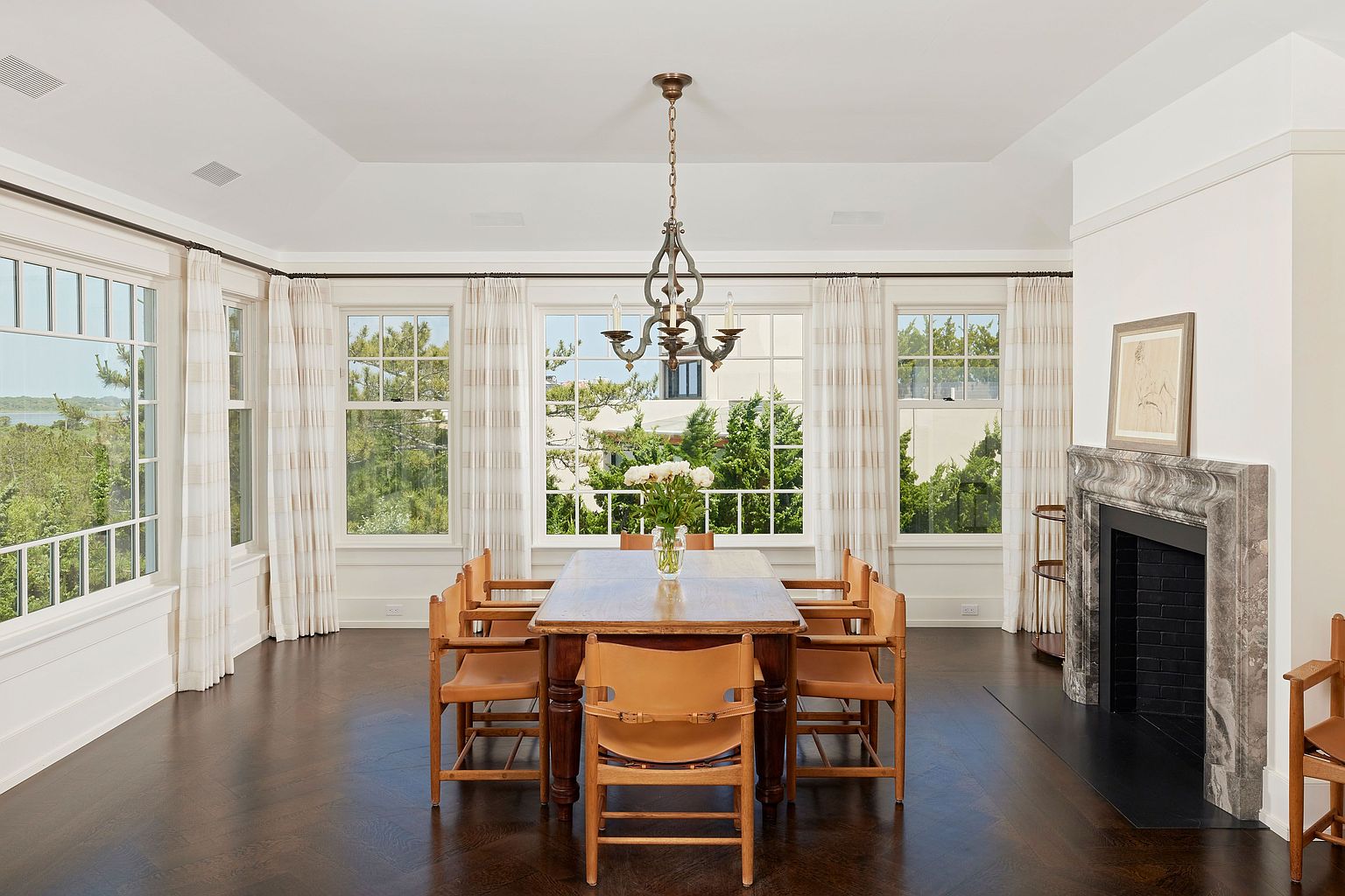 This is an interior shot of a dining room featuring a wooden dining table surrounded by six leather and wood chairs. A chandelier hangs above the table, and large windows with striped curtains provide natural light and views of the outdoors. A fireplace with a marble surround is visible on the right side of the room, adding a touch of elegance.
