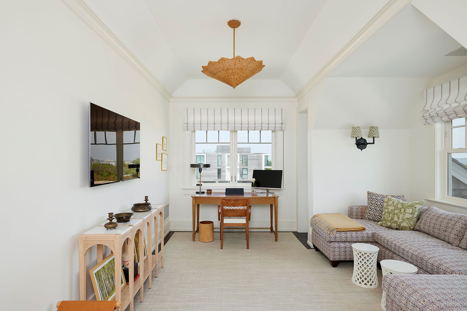 This is an interior shot of a bright and airy home office. The room features a desk with a computer, a console table with decorative items, and a comfortable seating area with a patterned sofa. The space is well-lit with natural light from a window and a unique pendant light fixture, creating a welcoming and productive atmosphere.