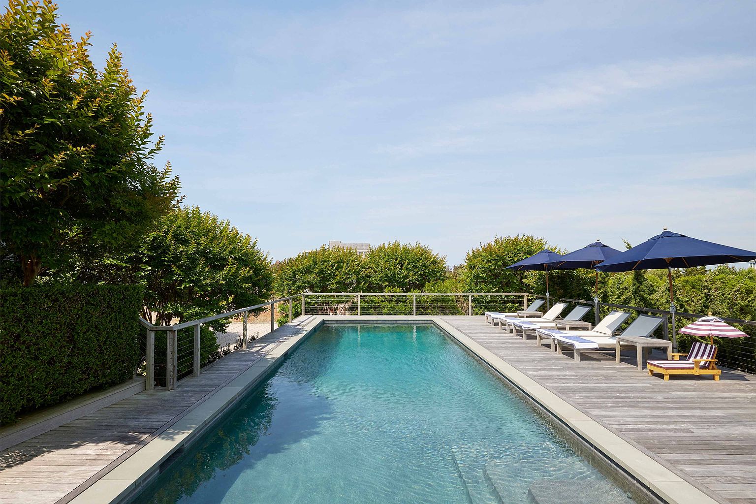 This image showcases a rectangular swimming pool surrounded by a wooden deck, complete with lounge chairs and umbrellas. The pool's water is a clear turquoise, reflecting the sky above. Lush greenery surrounds the area, providing privacy and a serene atmosphere, making it an ideal outdoor relaxation space.