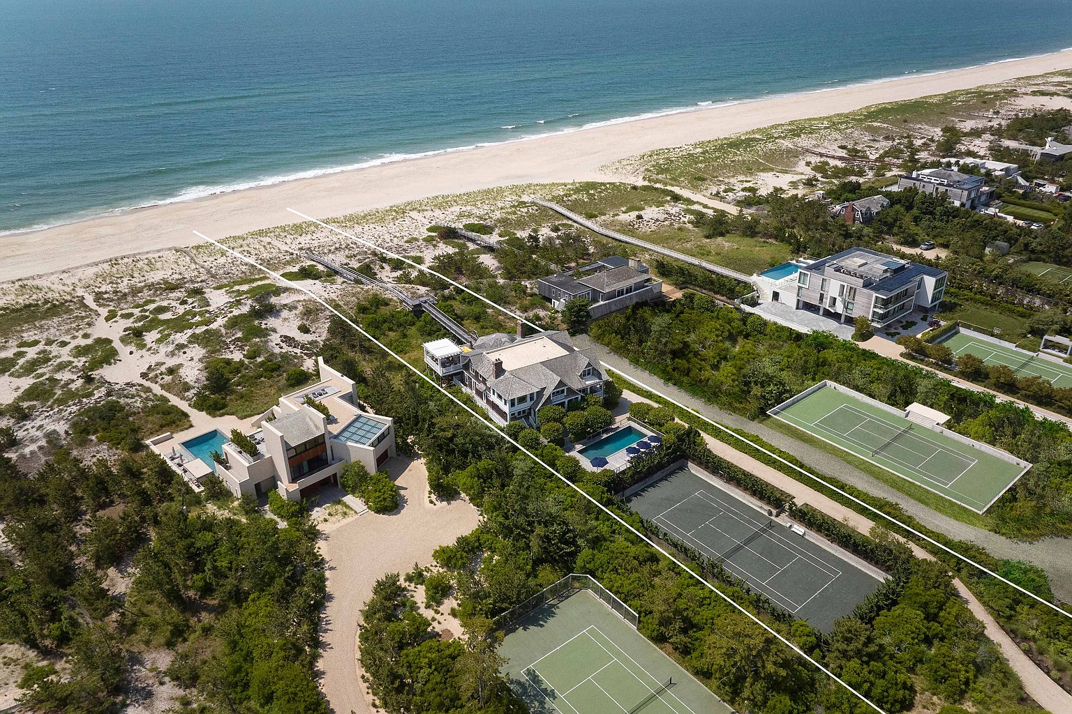 This aerial shot showcases a luxurious beachfront property featuring multiple modern homes, each with unique architectural designs. The properties include amenities such as swimming pools, tennis courts, and private beach access, all nestled within lush greenery. The overall impression is one of exclusivity and high-end coastal living.