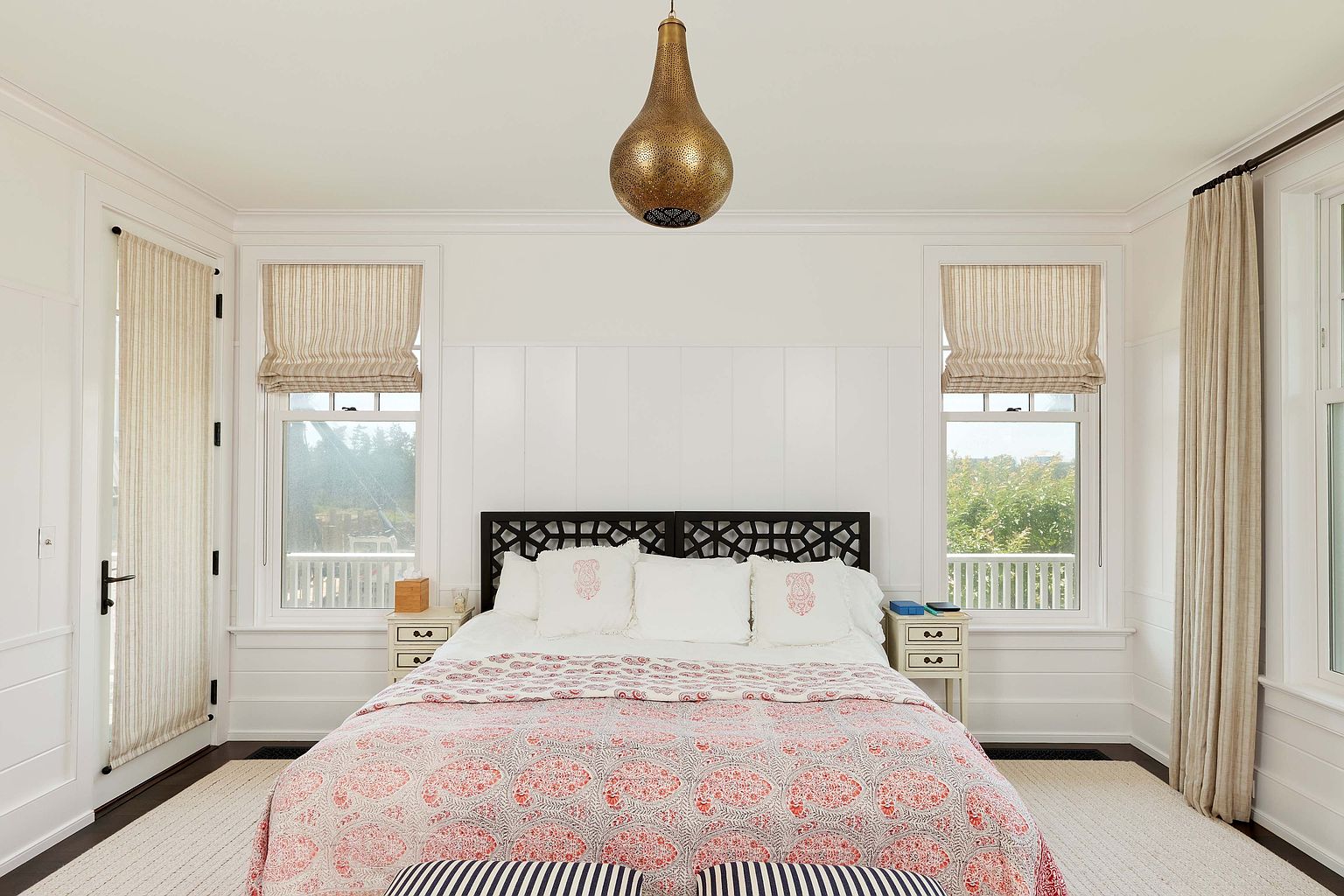 This is a well-lit primary bedroom featuring a bed with a patterned quilt and decorative pillows, flanked by matching nightstands. The room has a neutral color palette with white walls and trim, complemented by beige window treatments and a striking gold pendant light fixture. The overall impression is one of calm and sophisticated comfort.