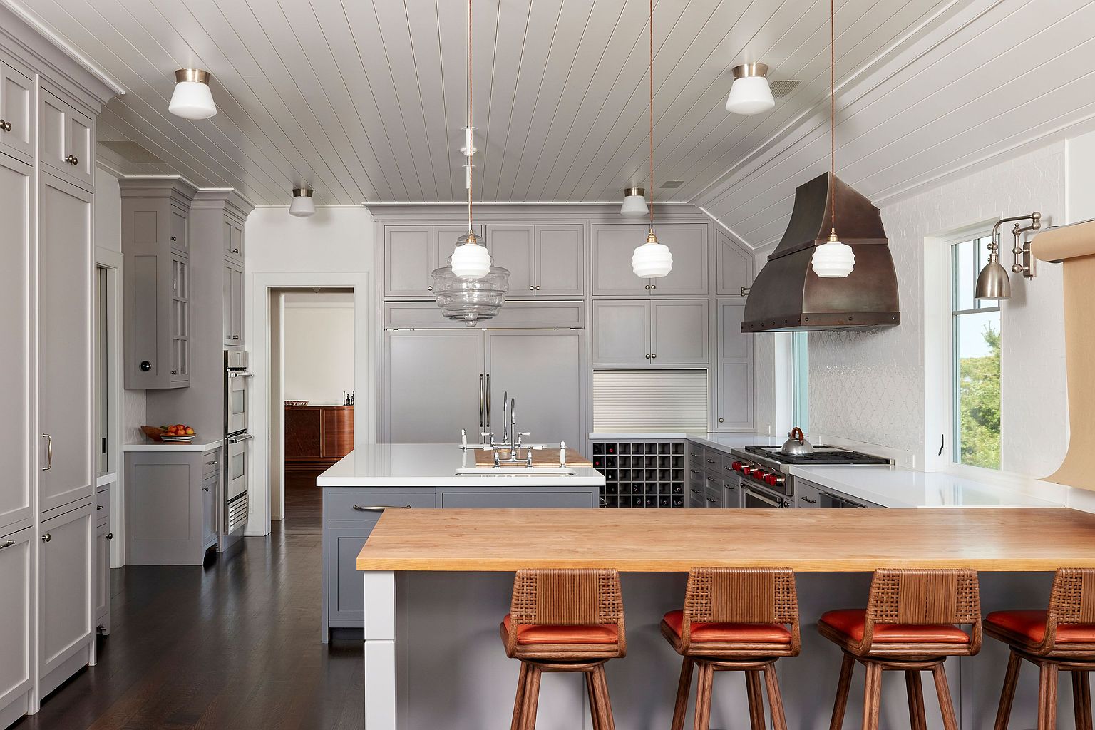 This is a well-lit kitchen featuring gray cabinetry, stainless steel appliances, and a large island with a white countertop and a wooden extension for seating. Pendant lights hang above the island, and a window provides natural light. The kitchen has a clean and modern aesthetic with a touch of traditional charm.