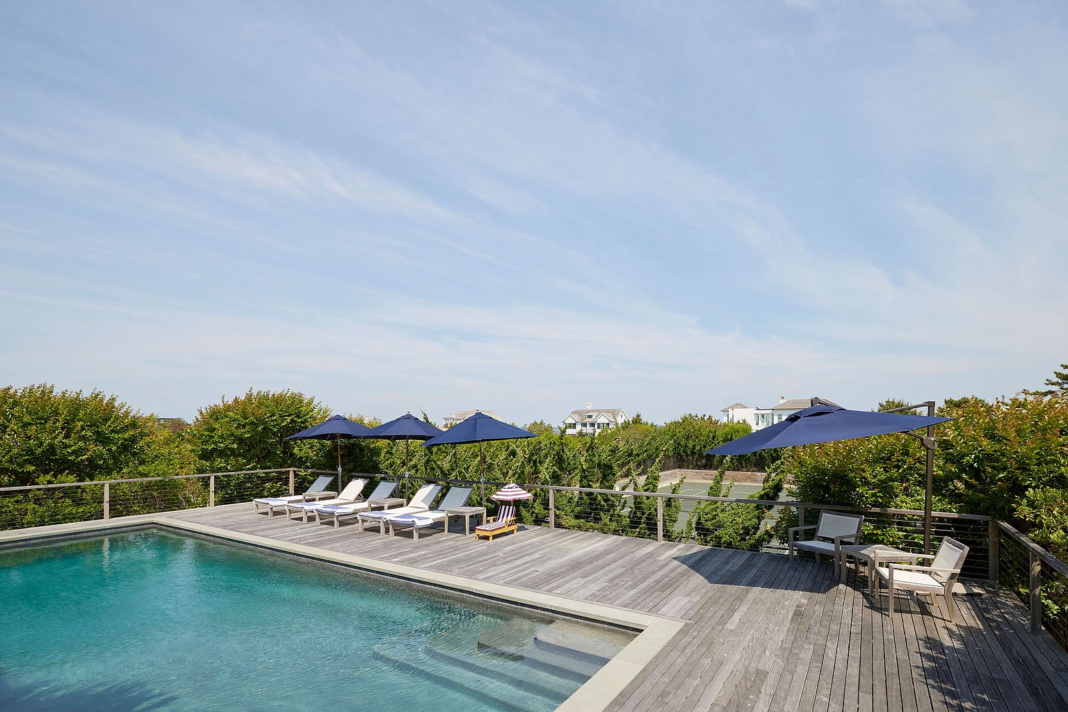 This image showcases a luxurious outdoor pool area, complete with a wooden deck, lounge chairs, and umbrellas for shade. The pool's clear water invites relaxation, while the surrounding greenery provides privacy and a sense of tranquility. The scene suggests a high-end property perfect for leisure and entertaining.