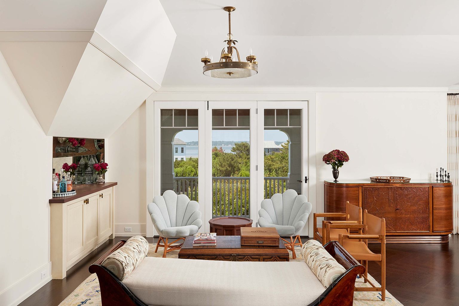 This is an interior shot of a living room featuring a daybed, two shell-backed chairs, and a wooden coffee table. The room has a light and airy feel, with natural light streaming in through the French doors that lead to a balcony. A wooden cabinet and a bar area are also visible, adding to the room's character and functionality.