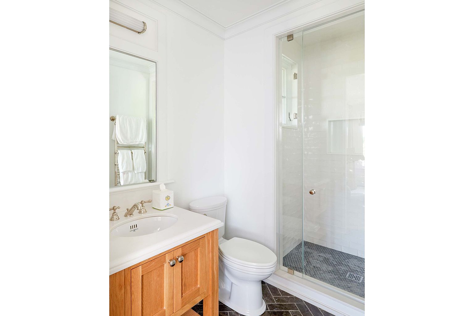 This is a well-lit bathroom featuring a wooden vanity with a white countertop and a sink. A toilet is positioned next to the vanity, and a glass-enclosed shower with white tiled walls and a dark tiled floor is visible. The bathroom has a clean and classic design with white walls and trim.