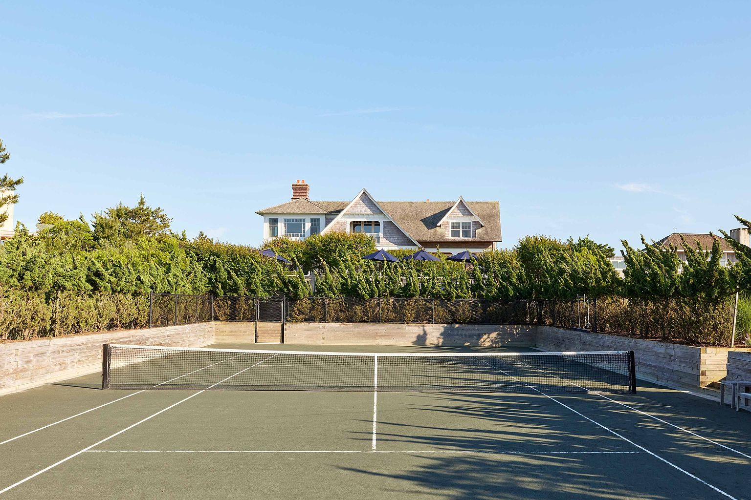 The image showcases a well-maintained tennis court with a dark green surface and crisp white lines, enclosed by a wooden retaining wall and a black fence. Lush green hedges line the fence, providing privacy and a sense of seclusion, while a charming house peeks out from behind the greenery. The scene is bathed in bright sunlight under a clear blue sky, creating an inviting and upscale atmosphere.